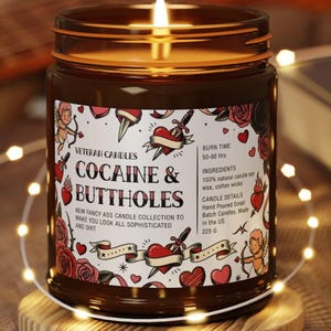 Puede incluir: Una vela encendida en un frasco de vidrio marr&oacute;n con una etiqueta que dice "COCAINE & BUTTHOLES" con gr&aacute;ficos estilo tatuaje. La vela est&aacute; sobre un posavasos de madera, rodeada de luces de cadena. La etiqueta tambi&eacute;n dice "NEW FANCY ASS CANDLE COLLECTION TO MAKE YOU LOOK ALL SOPHISTICATED AND SHIT."