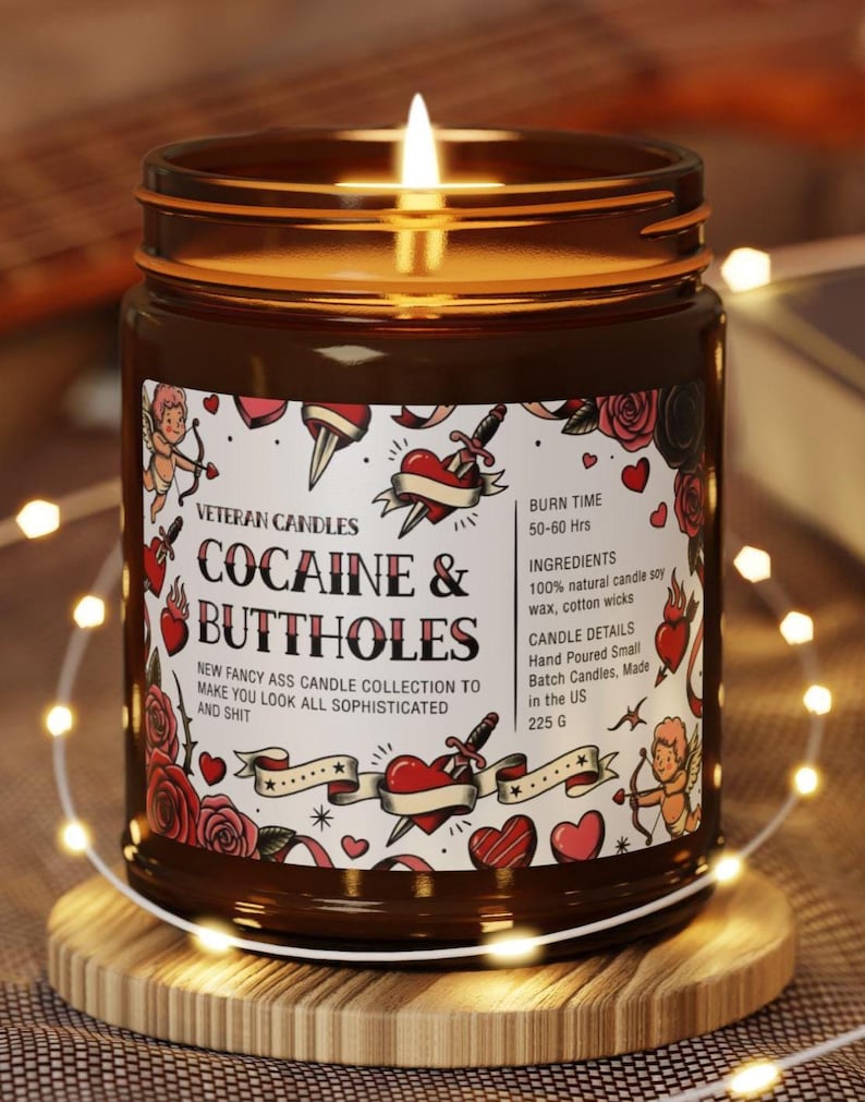 Puede incluir: Una vela encendida en un frasco de vidrio marr&oacute;n con una etiqueta que dice "COCAINE & BUTTHOLES" con gr&aacute;ficos estilo tatuaje. La vela est&aacute; sobre un posavasos de madera, rodeada de luces de cadena. La etiqueta tambi&eacute;n dice "NEW FANCY ASS CANDLE COLLECTION TO MAKE YOU LOOK ALL SOPHISTICATED AND SHIT."