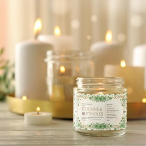 Op de afbeelding: Een brandende kaars in een glazen pot met het opschrift "COCAINE & BUTTHOLES" staat centraal. Verschillende andere brandende kaarsen van verschillende groottes staan op de achtergrond. De kaarsen staan op een houten oppervlak.