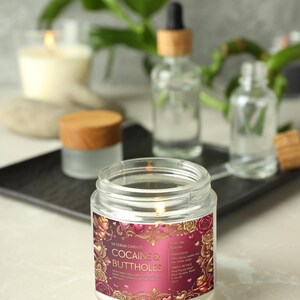 Peut inclure: Une bougie en pot de verre allum&eacute;e avec une &eacute;tiquette qui dit "COCAINE & BUTTHOLES" en lettres dor&eacute;es. L'&eacute;tiquette a un fond bordeaux avec des motifs floraux et de c&oelig;urs. D'autres bouteilles en verre et une bougie sont en arri&egrave;re-plan.
