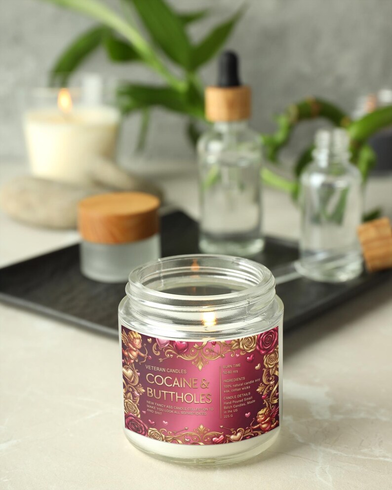 Peut inclure: Une bougie en pot de verre allum&eacute;e avec une &eacute;tiquette qui dit "COCAINE & BUTTHOLES" en lettres dor&eacute;es. L'&eacute;tiquette a un fond bordeaux avec des motifs floraux et de c&oelig;urs. D'autres bouteilles en verre et une bougie sont en arri&egrave;re-plan.