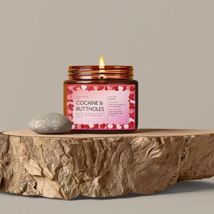 May include: A lit candle in a glass jar with a copper lid, labeled "COCAINE & BUTTHOLES" with a heart-shaped border. A small, gray stone sits next to the candle on a wooden slab.
