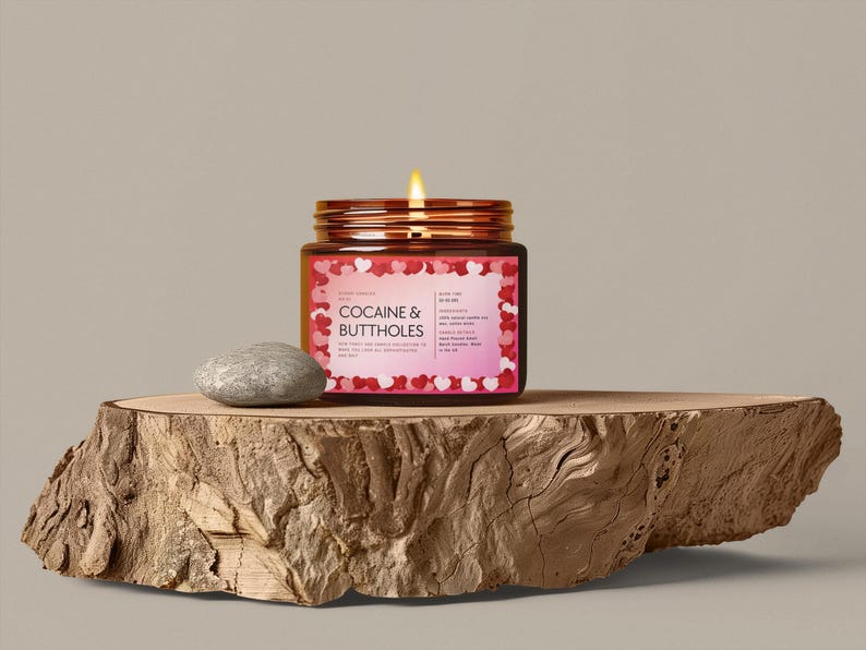 May include: A lit candle in a glass jar with a copper lid, labeled "COCAINE & BUTTHOLES" with a heart-shaped border. A small, gray stone sits next to the candle on a wooden slab.