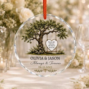 May include: A circular glass ornament with a red ribbon, featuring a printed illustration of a tree with a heart-shaped swing. The text reads: Grow old with me, the best is yet to be. OLIVIA & JASON, Always & Forever, and the date 06.15.2025.