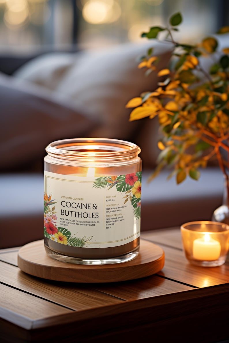 May include: A lit candle in a glass jar with the words "COCAINE & BUTTHOLES" on the label, decorated with tropical flowers. The candle sits on a wooden coaster, with a smaller candle in a glass holder nearby.