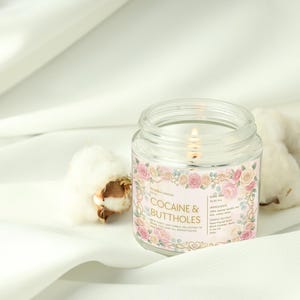 Puede incluir: Una vela encendida en un tarro de vidrio transparente con una etiqueta rosa con dise&ntilde;os florales y el texto "COCAINE & BUTTHOLES". La vela est&aacute; sobre una superficie de tela blanca junto a capullos de algod&oacute;n. La etiqueta de la vela tambi&eacute;n enumera los ingredientes.