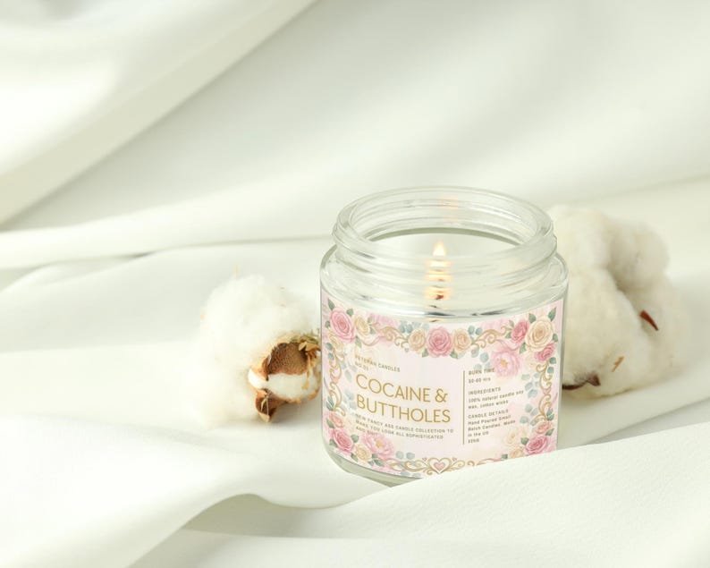Puede incluir: Una vela encendida en un tarro de vidrio transparente con una etiqueta rosa con dise&ntilde;os florales y el texto "COCAINE & BUTTHOLES". La vela est&aacute; sobre una superficie de tela blanca junto a capullos de algod&oacute;n. La etiqueta de la vela tambi&eacute;n enumera los ingredientes.