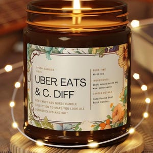 May include: A lit brown glass jar candle with a floral label that reads "UBER EATS & C. DIFF". The candle sits on a wooden coaster, surrounded by string lights. The label also states "NEW FANCY ASS NURSE CANDLE COLLECTION TO MAKE YOU LOOK ALL SOPHISTICATED AND SHIT."