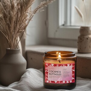 May include: A lit candle in a brown glass jar with a label that reads "COCAINE & BUTTHOLES". The label has a pink background with red and pink hearts. The candle is on a white cloth.