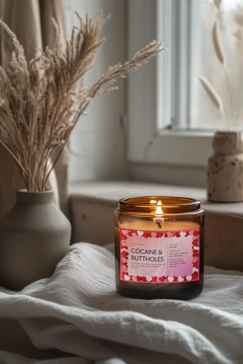 May include: A lit candle in a brown glass jar with a label that reads "COCAINE & BUTTHOLES". The label has a pink background with red and pink hearts. The candle is on a white cloth.