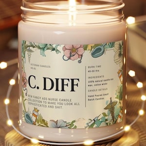 May include: A lit, pink candle in a glass jar with a label featuring floral and medical illustrations. The label reads "C. DIFF" and "New Fancy Ass Nurse Candle Collection". The candle sits on a wooden coaster.