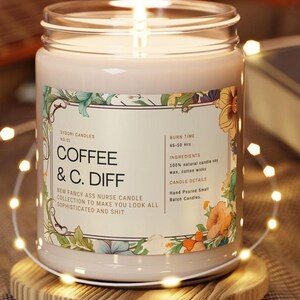 May include: A lit, cream-coloured candle in a glass jar with a floral label. The label reads "COFFEE & C. DIFF" and "NEW FANCY ASS NURSE CANDLE COLLECTION". The candle sits on a wooden coaster, surrounded by string lights.