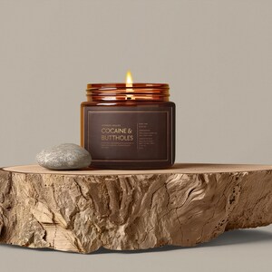 May include: A lit candle in a brown glass jar with the text "COCAINE & BUTTHOLES" on a wooden slab. A small, gray stone sits next to the jar. The candle has a copper-colored lid.