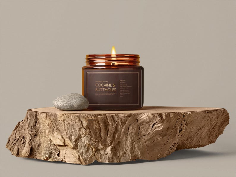 May include: A lit candle in a brown glass jar with the text "COCAINE & BUTTHOLES" on a wooden slab. A small, gray stone sits next to the jar. The candle has a copper-colored lid.