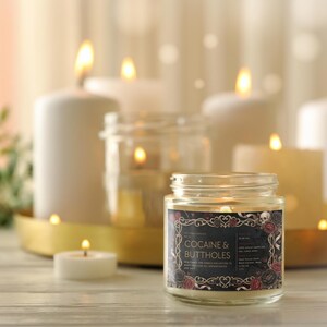 May include: A lit jar candle with the text "COCAINE & BUTTHOLES" on a black label with floral and skull accents. Several other lit candles of varying sizes are in the background, along with a gold tray.