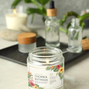 May include: A lit, clear glass jar candle with a label that reads "COCAINE & BUTTHOLES" with a floral design. Other glass bottles and a candle are in the background. The candle is part of the "NEW FANCY AND CANDLE COLLECTION TO MAKE YOU LOOK ALL SOPHISTICATED AND SASSY."