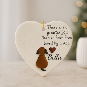Puede incluir: Adorno en forma de coraz&oacute;n crema con una ilustraci&oacute;n de perro y el texto "No hay mayor alegr&iacute;a que haber sido amado por un perro." El perro es marr&oacute;n con orejas y cola negras. El nombre "Bella" est&aacute; debajo.