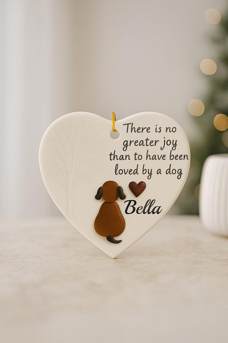 Puede incluir: Adorno en forma de coraz&oacute;n crema con una ilustraci&oacute;n de perro y el texto "No hay mayor alegr&iacute;a que haber sido amado por un perro." El perro es marr&oacute;n con orejas y cola negras. El nombre "Bella" est&aacute; debajo.