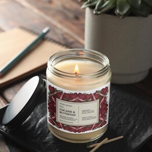 May include: A lit candle in a glass jar with a label that reads "Cocaine & Buttholes." The label has a red and white heart design. The candle sits on a black tray with a lid next to it. A notebook, pencil, and plant are in the background.