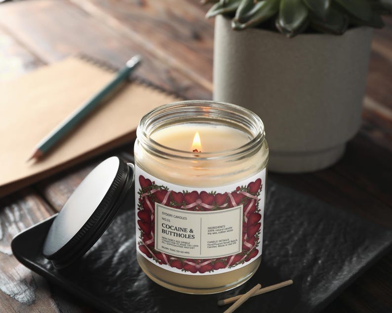 May include: A lit candle in a glass jar with a label that reads "Cocaine & Buttholes." The label has a red and white heart design. The candle sits on a black tray with a lid next to it. A notebook, pencil, and plant are in the background.