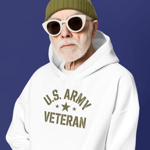 Puede incluir: Una sudadera con capucha blanca con el texto "U.S. ARMY VETERAN" en verde oliva. La persona lleva un gorro verde y gafas de sol con montura blanca. El fondo es azul liso.