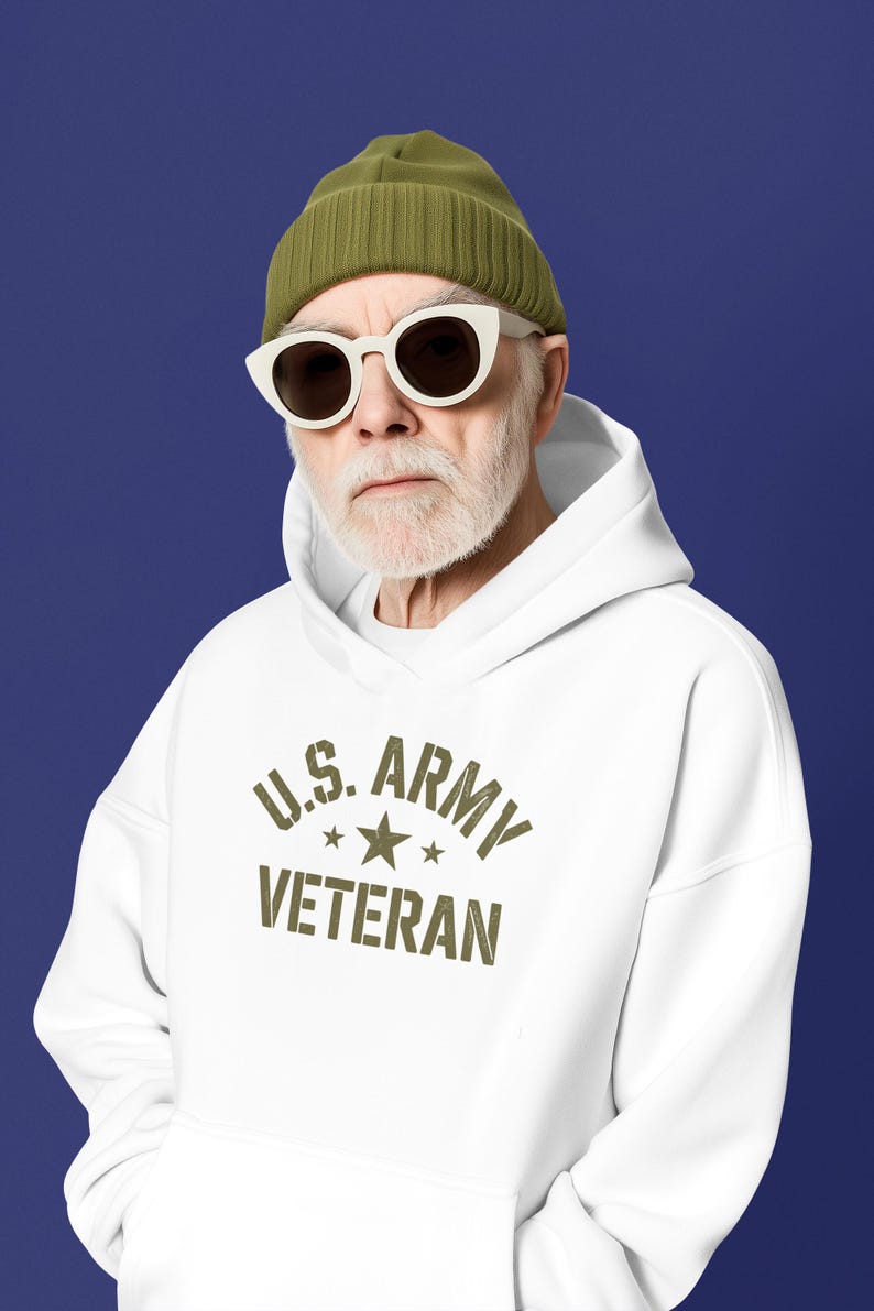Puede incluir: Una sudadera con capucha blanca con el texto "U.S. ARMY VETERAN" en verde oliva. La persona lleva un gorro verde y gafas de sol con montura blanca. El fondo es azul liso.