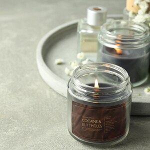 May include: A lit, clear glass jar candle with a brown label that reads "COCAINE & BUTTHOLES". The candle is surrounded by other candles, a perfume bottle, and white flowers on a gray tray.