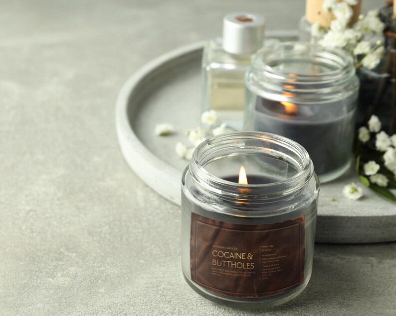 May include: A lit, clear glass jar candle with a brown label that reads "COCAINE & BUTTHOLES". The candle is surrounded by other candles, a perfume bottle, and white flowers on a gray tray.