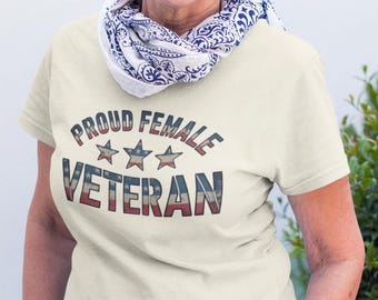 Puede incluir: Camiseta color crema con el texto "PROUD FEMALE VETERAN" en rojo, blanco y azul. El texto está sobre tres estrellas. La persona lleva una bufanda con estampado azul y blanco.