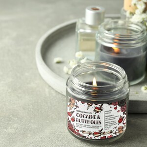 Peut inclure: Une bougie allum&eacute;e de couleur fonc&eacute;e dans un pot en verre transparent avec une &eacute;tiquette qui dit "Cocaine & Buttholes". L'&eacute;tiquette pr&eacute;sente des illustrations de style tatouage de c&oelig;urs, de roses et de ch&eacute;rubins. D'autres bougies et objets d&eacute;coratifs sont en arri&egrave;re-plan.