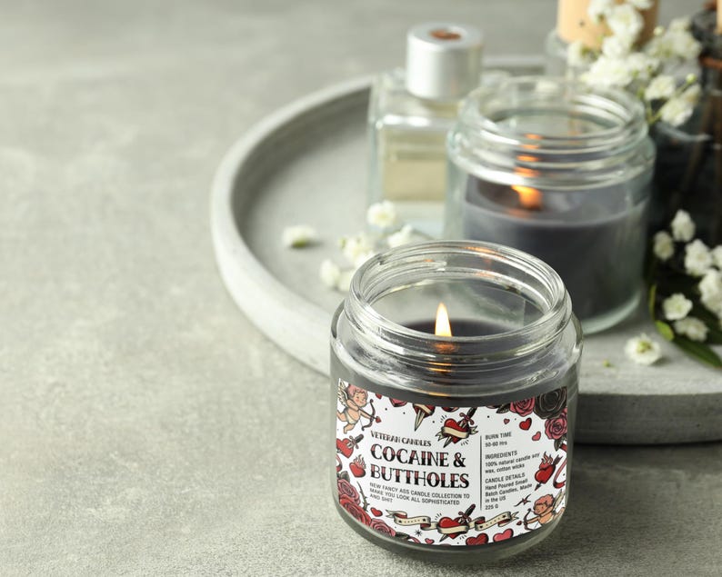 Peut inclure: Une bougie allum&eacute;e de couleur fonc&eacute;e dans un pot en verre transparent avec une &eacute;tiquette qui dit "Cocaine & Buttholes". L'&eacute;tiquette pr&eacute;sente des illustrations de style tatouage de c&oelig;urs, de roses et de ch&eacute;rubins. D'autres bougies et objets d&eacute;coratifs sont en arri&egrave;re-plan.