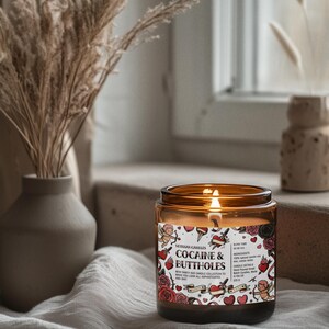 Puede incluir: Un tarro de vela de vidrio &aacute;mbar encendido con una etiqueta que dice "COCAINE & BUTTHOLES" con un dise&ntilde;o de estilo tatuaje. La vela est&aacute; sobre una tela blanca, con un jarr&oacute;n de plantas secas en el fondo.