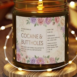 May include: A lit candle in a glass jar with a gold lid, resting on a wooden coaster. The label reads "COCAINE & BUTTHOLES" with floral accents. The candle is part of the "NEW FANCY ASS CANDLE COLLECTION".