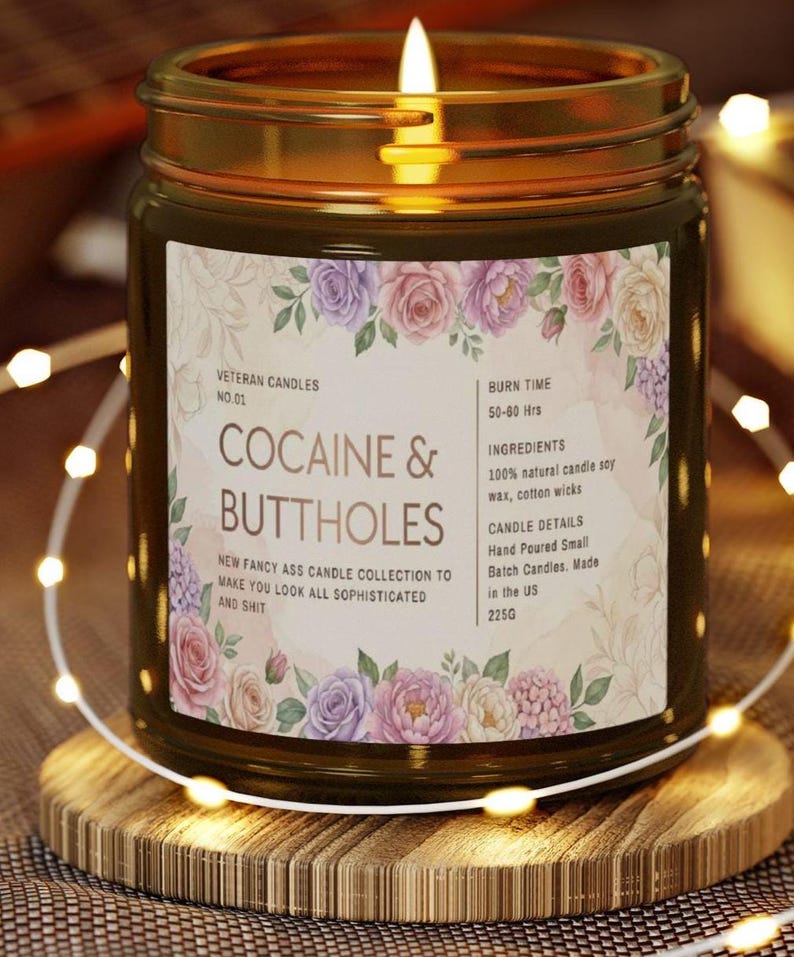 May include: A lit candle in a glass jar with a gold lid, resting on a wooden coaster. The label reads "COCAINE & BUTTHOLES" with floral accents. The candle is part of the "NEW FANCY ASS CANDLE COLLECTION".