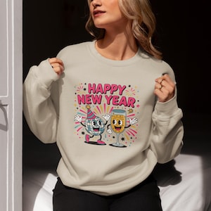 May include: A light beige sweatshirt with a colorful graphic that says "HAPPY NEW YEAR". The graphic features a disco ball character wearing a party hat and a champagne glass character, both with smiling faces and outstretched arms.