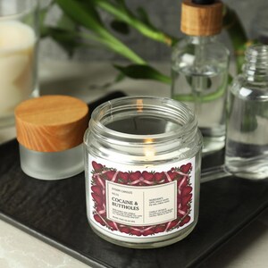 Puede incluir: Una vela encendida en un tarro de cristal transparente con tapa de madera, etiquetada "COCAINE & BUTTHOLES" con un borde de coraz&oacute;n rojo. Otras botellas de vidrio y una vela encendida est&aacute;n en el fondo, todo sobre una bandeja negra.