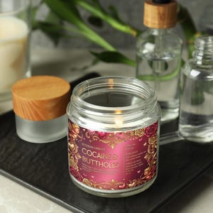 Peut inclure: Une bougie en pot de verre allum&eacute;e avec un couvercle en bois, portant l'inscription "COCAINE'S BUTTHOLES". La bougie est pos&eacute;e sur un plateau noir, avec d'autres flacons en verre et une bougie allum&eacute;e en arri&egrave;re-plan.