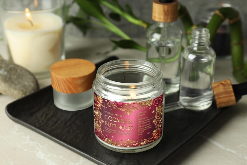 Peut inclure: Une bougie en pot de verre allum&eacute;e avec un couvercle en bois, portant l'inscription "COCAINE'S BUTTHOLES". La bougie est pos&eacute;e sur un plateau noir, avec d'autres flacons en verre et une bougie allum&eacute;e en arri&egrave;re-plan.