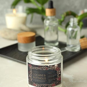 May include: A lit, clear glass jar candle with a label that reads "Cocaine & Buttholes." The label has a decorative border with roses and skulls. Other glass bottles and a small jar with a wooden lid are in the background.
