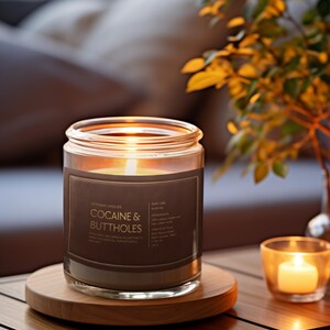 May include: A lit candle in a glass jar with a brown label that reads "COCAINE & BUTTHOLES" sits on a wooden coaster. A smaller candle in a glass votive and a vase of greenery are also on the table. The candle is part of the "VINTAGE CANDLES" collection.