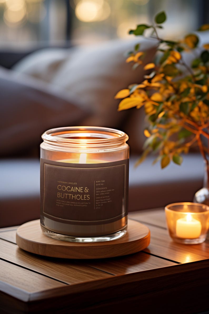 May include: A lit candle in a glass jar with a brown label that reads "COCAINE & BUTTHOLES" sits on a wooden coaster. A smaller candle in a glass votive and a vase of greenery are also on the table. The candle is part of the "VINTAGE CANDLES" collection.