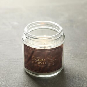 May include: A lit, clear glass jar candle with a brown label. The label reads "COCAINE & BUTTHOLES" in white text. The candle sits on a gray surface. The candle is a home decor item.
