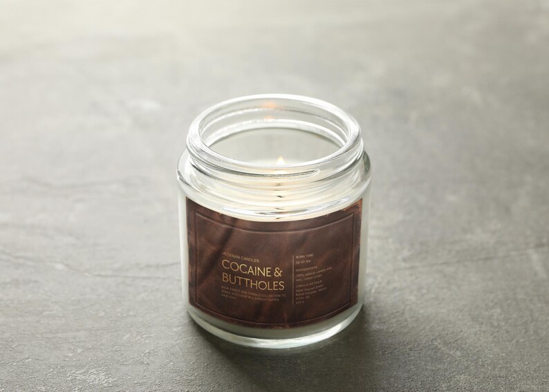May include: A lit, clear glass jar candle with a brown label. The label reads "COCAINE & BUTTHOLES" in white text. The candle sits on a gray surface. The candle is a home decor item.