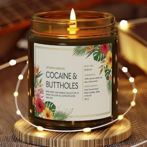May include: A lit candle in a glass jar with a gold rim, sitting on a wooden coaster. The label reads "COCAINE & BUTTHOLES" with floral accents. The candle is part of the "NEW FANCY ASS CANDLE COLLECTION". The candle weighs 225 grams.