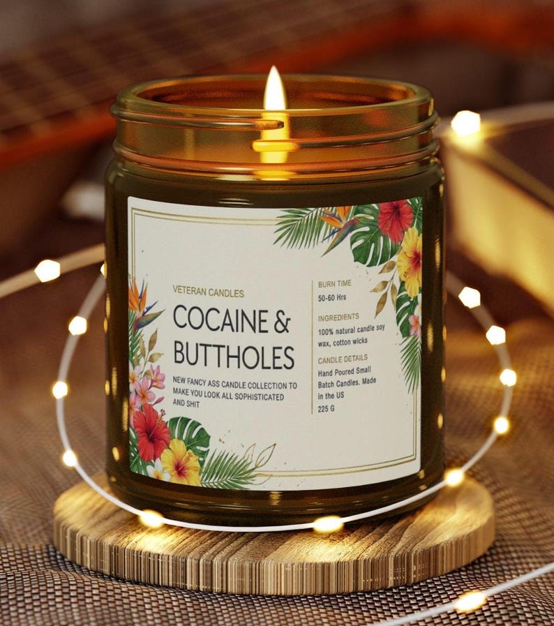 May include: A lit candle in a glass jar with a gold rim, sitting on a wooden coaster. The label reads "COCAINE & BUTTHOLES" with floral accents. The candle is part of the "NEW FANCY ASS CANDLE COLLECTION". The candle weighs 225 grams.
