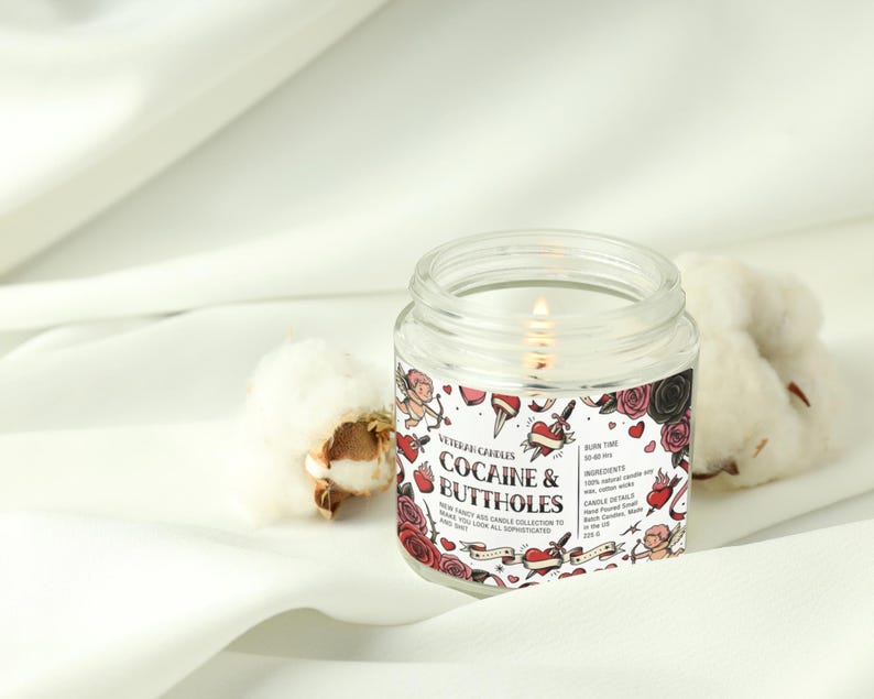 Peut inclure: Une bougie en pot de verre transparent avec une m&egrave;che allum&eacute;e, &eacute;tiquet&eacute;e "Cocaine & Buttholes". L'&eacute;tiquette pr&eacute;sente des illustrations de style tatouage de roses, de coeurs et de ch&eacute;rubins. La bougie est pos&eacute;e sur un tissu blanc avec des cotons.