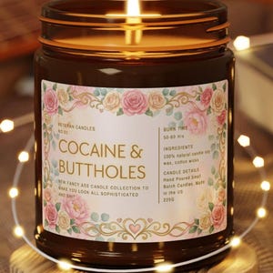 Pu&ograve; includere: Una candela accesa in un barattolo di vetro marrone con un'etichetta floreale che recita "COCAINE & BUTTHOLES". L'etichetta include il testo "Veteran Candles" e "New Fancy Ass Candle Collection". La candela &egrave; su un sottobicchiere di legno, circondata da luci a catena.