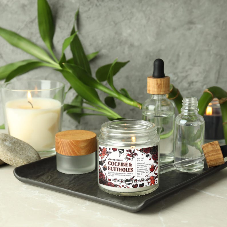 Może przedstawiać: Zapalone świeca w szklanym słoiku z etykietą "Cocaine & Buttholes" stoi na czarnej tacy z innymi świecami i butelkami. Etykieta ma czerwono-białą grafikę. Łodygi i liście bambusa w tle.