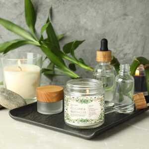 Op de afbeelding: Een brandende kaars in een glazen pot met de tekst "COCAINE & BUTTHOLES" op een etiket, weergegeven met andere kaarsen, glazen flessen en bamboe op een zwarte tray. De kaars heeft een houten deksel en is omgeven door groene bladeren.