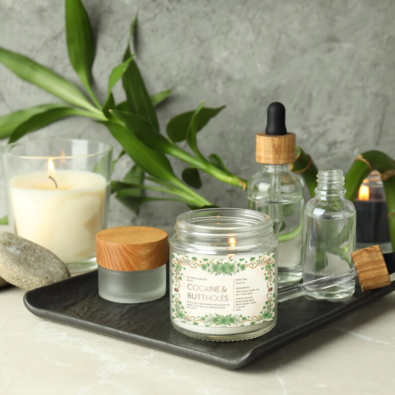 Op de afbeelding: Een brandende kaars in een glazen pot met de tekst "COCAINE & BUTTHOLES" op een etiket, weergegeven met andere kaarsen, glazen flessen en bamboe op een zwarte tray. De kaars heeft een houten deksel en is omgeven door groene bladeren.